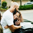 A father embracing his trans teenager, standing beside the kid's mother, in front of a car outside.