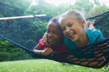 A Black girl and white girl swing on a hammock on their stomachs, laughing together.