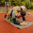 A man doing planks outside on a basketball court with his child on his back.