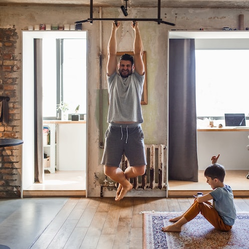 A man does a chin up bodyweight back exercise at home while his son sits with him.