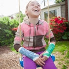 A girl with two broken arms in casts smiles and laughs while sitting on a swing outdoors.