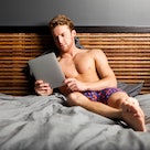 A man in his underwear on his bed, looking at his tablet.
