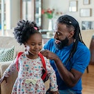 A dad helps his daughter get ready for Kindergarten by putting on her backpack at home.