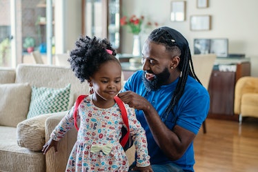 A dad helps his daughter get ready for Kindergarten by putting on her backpack at home.