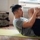 A man stretches on his back on a yoga mat at home, pulling his knee to his chest.