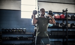 A man lifting dumbbells, doing a standing shoulder press, at the gym for strength training.