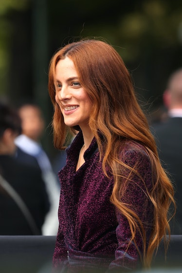 Riley Keough attends a fashion show. Her red hair falls down her shoulders and she wears a dark purp...