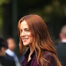 Riley Keough attends a fashion show. Her red hair falls down her shoulders and she wears a dark purp...