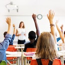 School kids in classroom raising their hands.