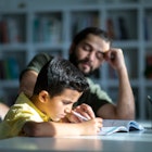 A dad leaning on his elbow helps his high-achieving son who is writing, working on a school assignme...