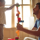 A dad helps his child build a tall tower out of blocks.