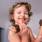 A baby holds a chocolate ice cream cone in each hand and has chocolate ice cream smeared on their fa...
